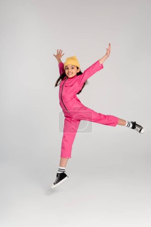 glückliches Mädchen mit Hut und stylischem rosa Outfit, das mit ausgestreckten Händen auf grau springt, viel Spaß