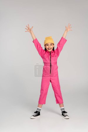 happy girl in beanie hat and pink outfit standing with outstretched hands on grey, have fun, style
