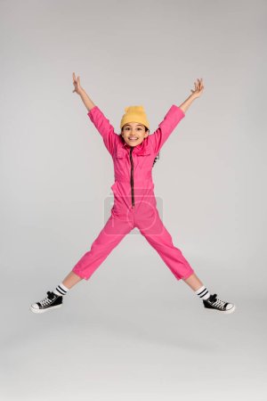 Téléchargez les photos : Fille excitée en bonnet à la mode chapeau et tenue rose sauter avec les mains tendues sur gris, amusez-vous - en image libre de droit