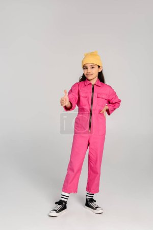 glückliches Kind mit gelbem Hut und rosafarbenem Outfit, das mit der Hand auf der Hüfte steht, wie ein Schild, grauer Hintergrund