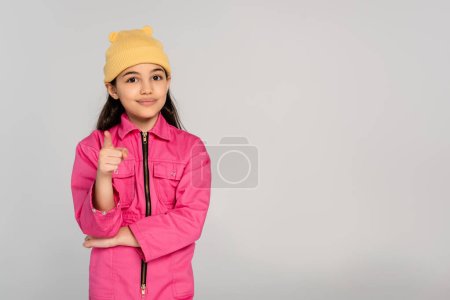 happy kid in yellow beanie hat and pink outfit pointing at camera on grey backdrop, stylish kid