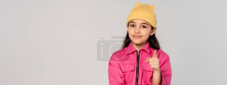 glückliches Kind mit gelbem Hut und rosa Outfit, das auf grauem Hintergrund in die Kamera zeigt, stylisch, Banner