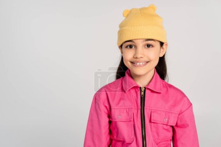 Photo for Happy girl in yellow beanie hat and pink outfit looking at camera on grey backdrop, stylish kid - Royalty Free Image