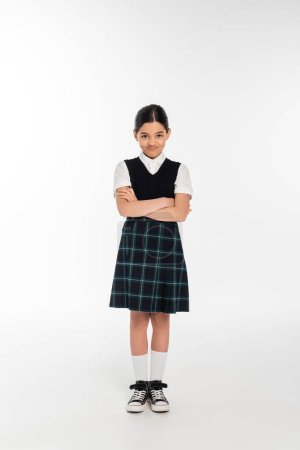 Foto de Longitud completa, escolar ofendido en uniforme mirando a la cámara y de pie con los brazos cruzados en blanco - Imagen libre de derechos