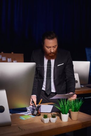 bearded and concentrated businessman working with documents near computer at night in office
