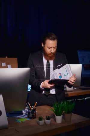 bearded businessman in suit working late and looking at documents with infographics in night office