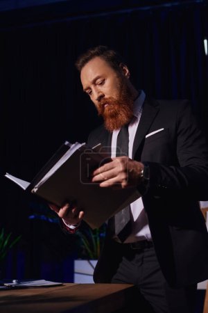 Foto de Hombre de negocios barbudo en ropa formal elegante que trabaja con documentos en la oficina por la noche, vista de ángulo bajo - Imagen libre de derechos