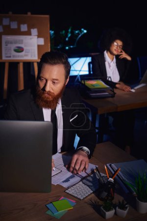 bearded man working with documents and computer near charming african american woman on background