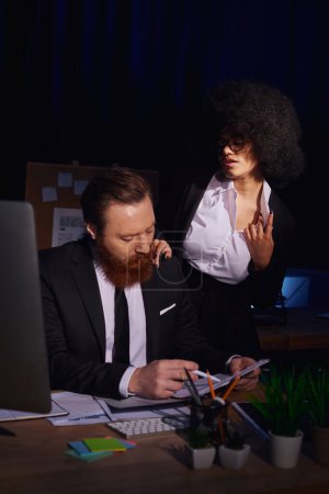 young african american woman touching shoulder and seducing serious businessman at night in office