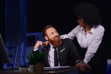 african american woman embracing happy bearded businessman in night office, love affair at work