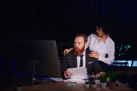 mujer afroamericana abrazando hombros de un colega barbudo trabajando con documentos en la oficina nocturna