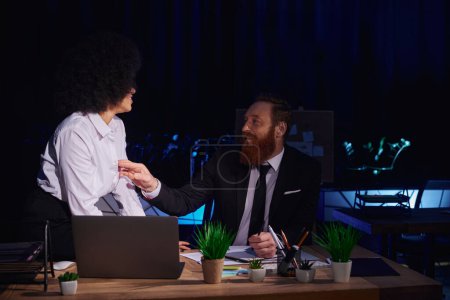 bearded businessman touching blouse of seductive african american woman near laptop in night office