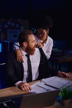 passionate african american woman undressing bearded businessman near documents and laptop in office