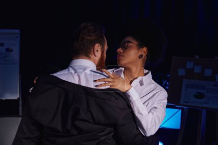 séduisante femme afro-américaine embrassant homme d'affaires barbu la nuit au bureau, flirtant au travail