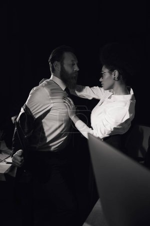 apasionada mujer afroamericana abrazando hombre de negocios en la noche en la oficina, foto en blanco y negro