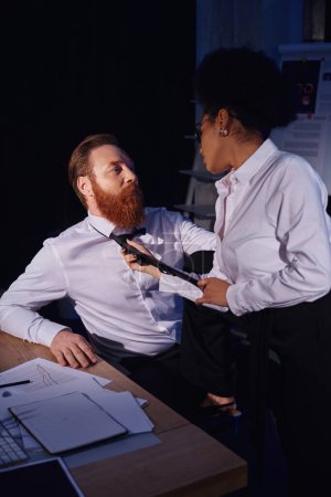 african american woman pulling tie of bearded businessman in night office, love affair at work