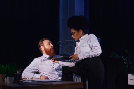 Photo for African american businesswoman pulling tie of bearded businessman at work desk in office, seduction - Royalty Free Image