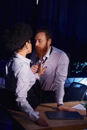 charismatic bearded businessman undressing near young african american woman in office at night