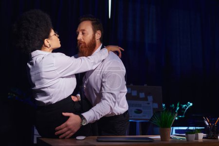 bearded businessman embracing hot african american woman sitting on desk, seduction in night office