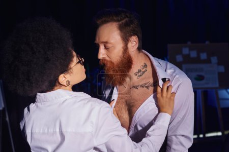 hot african american woman undressing bearded tattooed colleague in darkness, seduction in office