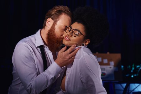 bearded businessman kissing smiling african american woman in eyeglasses, office romance at night