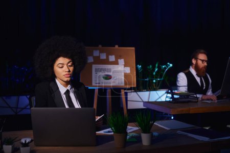 mujer afroamericana que trabaja en el ordenador portátil cerca de colega barbudo en el fondo en la oficina de noche