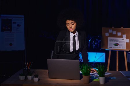 african american businesswoman in formal wear looking at laptop while working in office at night