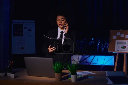 mujer de negocios afroamericana con portátil hablando en el teléfono inteligente cerca de la computadora portátil en la oficina por la noche