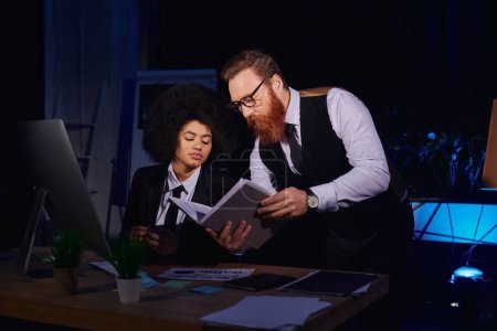 Photo for Bearded businessman showing notebook to african american secretary near computer in night office - Royalty Free Image