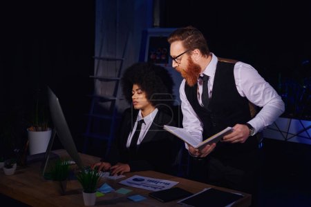 Afroamerikanerin tippt in der Nähe eines bärtigen Geschäftsmannes nachts mit Notizbuch am Computer