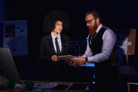 Photo for Businessman in eyeglasses showing documents to displeased african american secretary in night office - Royalty Free Image