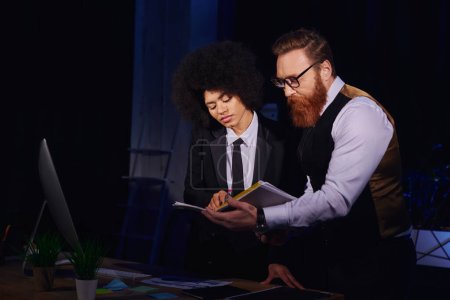 african american businesswoman and bearded colleague looking at documents in dark office, night work