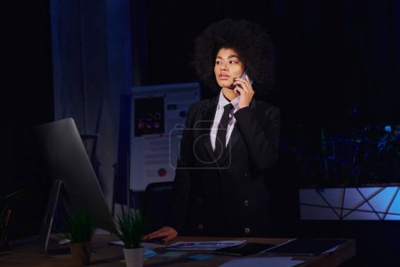 mujer de negocios afroamericana hablando en el teléfono inteligente cerca del monitor de la computadora en la oficina por la noche