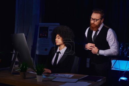 bärtiger Geschäftsmann schaut hübsche afrikanisch-amerikanische Sekretärin an, die im Nachtbüro am Computer tippt