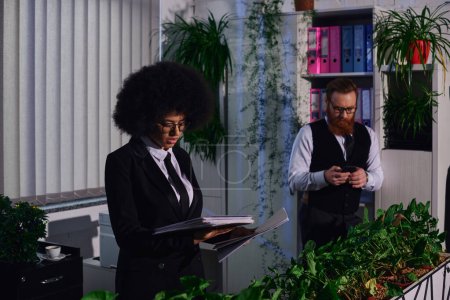 african american woman with documents near bearded colleague using smartphone at night in office
