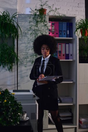 african american woman in formal wear standing with paper documents near copier at night in office