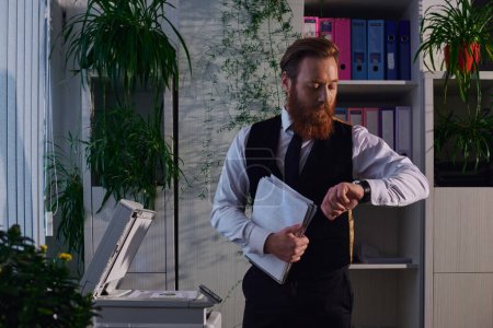 bearded businessman with papers looking at wristwatch near copier while working late in office