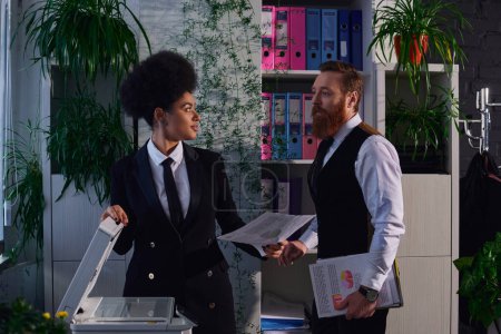 interracial colleagues with documents looking at each other near copier in office at night