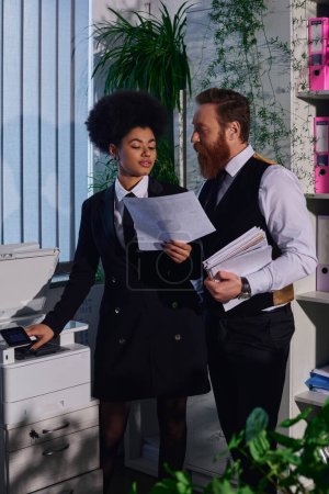 bearded businessman looking at african american secretary with document near copier in night office