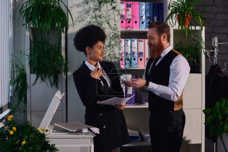 bearded businessman holding coffee cup and flirting with african american secretary in office