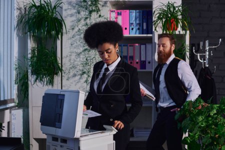 hombre de negocios barbudo mirando atractiva secretaria afroamericana cerca de fotocopiadora en la noche en la oficina