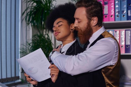 bearded businessman embracing african american woman with closed eyes and document in night office