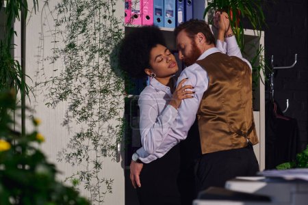 passionate interracial couple embracing near decorative plants in office at night, work romance