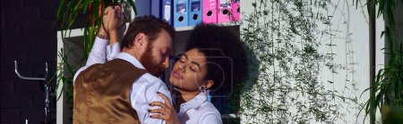 passionate interracial couple embracing near decorative plants in office at night, banner