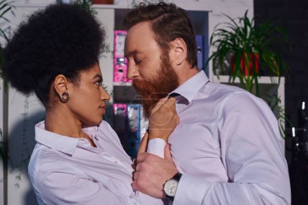 Téléchargez les photos : Vue latérale de séduisante femme afro-américaine flirtant avec homme d'affaires barbu dans le bureau de nuit - en image libre de droit
