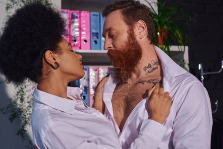 passionate african american woman undressing bearded tattooed businessman, romance in night office