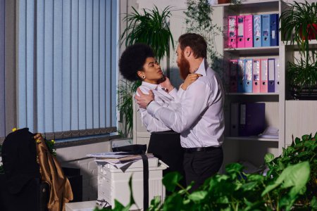 leidenschaftliches multiethnisches Paar, das sich nachts in der Nähe eines Kopierers im Büro umarmt, romantische Begegnung
