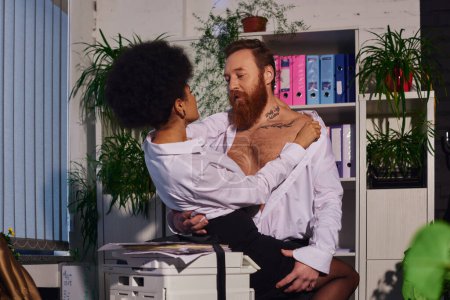 hombre de negocios tatuado barbudo abrazando a una joven afroamericana cerca de una fotocopiadora, pasión en el cargo