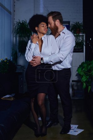 full length of stylish interracial couple in formal wear embracing in office, romantic encounter