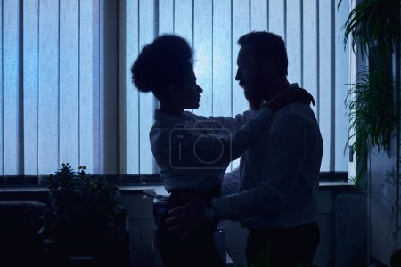 Foto de Siluetas oscuras de pareja multiétnica abrazándose en la oficina por la noche, encuentro romántico en el trabajo - Imagen libre de derechos